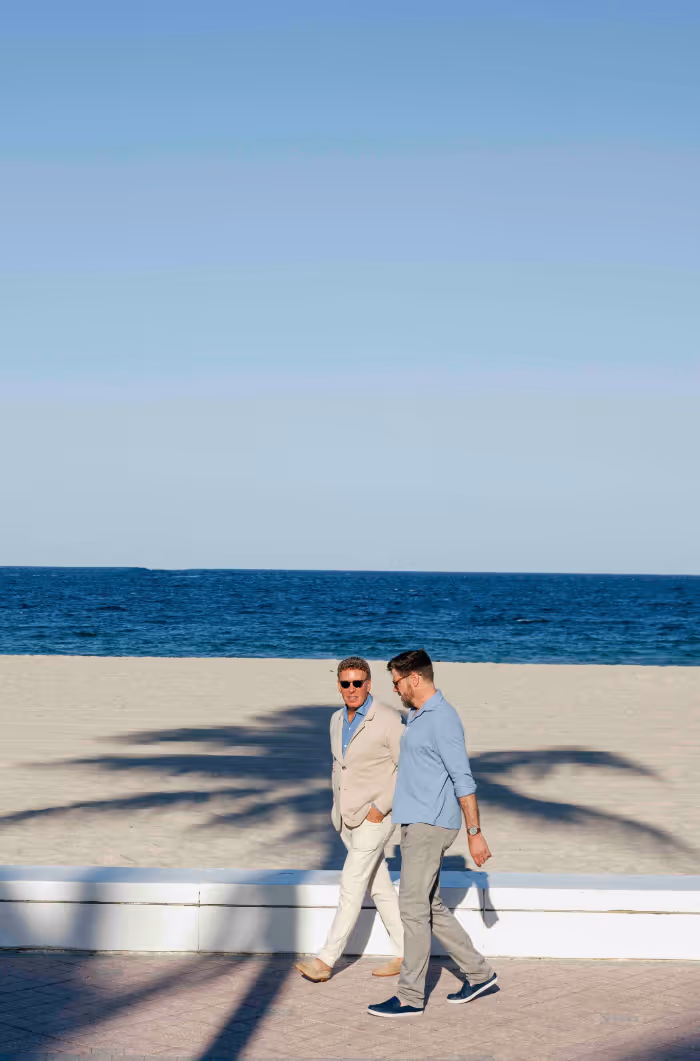 Two men walking and talking on a paved walkway by the beach with the ocean and clear sky in the background.