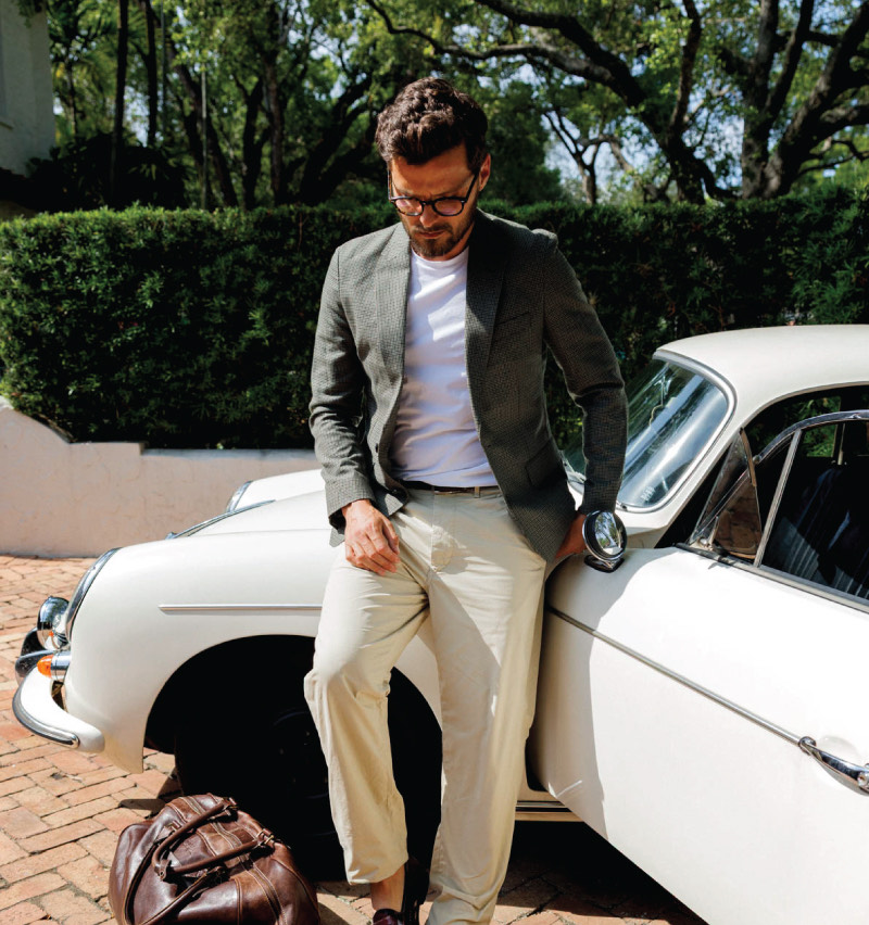 Man wearing glasses, a grey blazer, white shirt, and beige pants leaning against a white vintage car with a brown leather bag on the ground nearby.