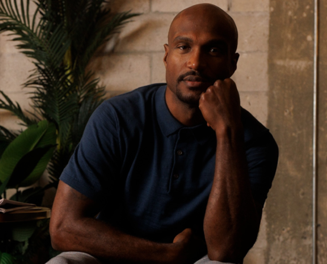 Portrait of a contemplative bald man with a beard resting his chin on his fist indoors near green plants and a textured wall.