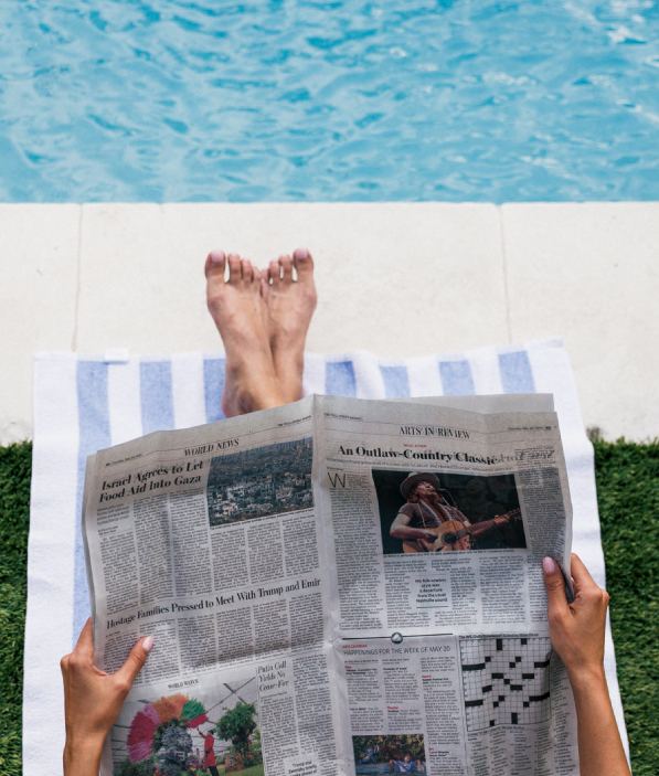 Person lounging by a pool on a striped towel, holding and reading a newspaper with visible articles and a crossword puzzle.