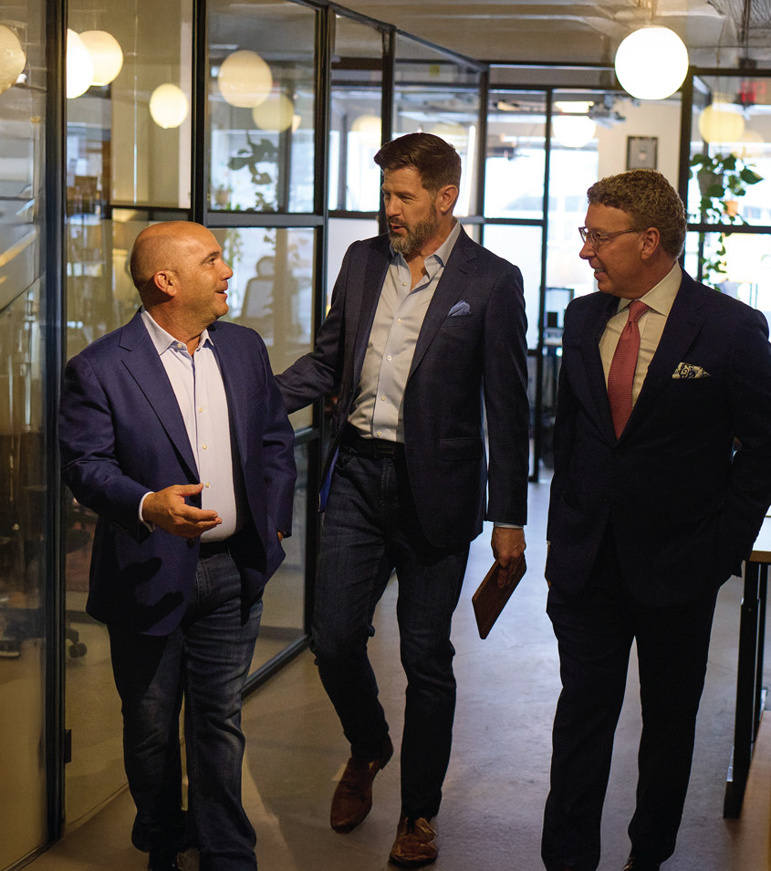 Wicho Hernandez, Jeremy Straub, and Harris Fishman in business attire walking and talking together in a modern office hallway with glass walls and round ceiling lights.