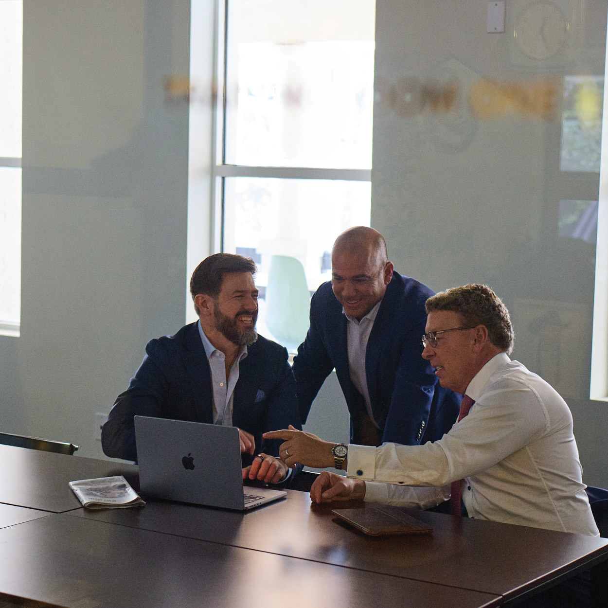 Three businessmen in formal attire gathered around a laptop, engaged in a discussion in a conference room.