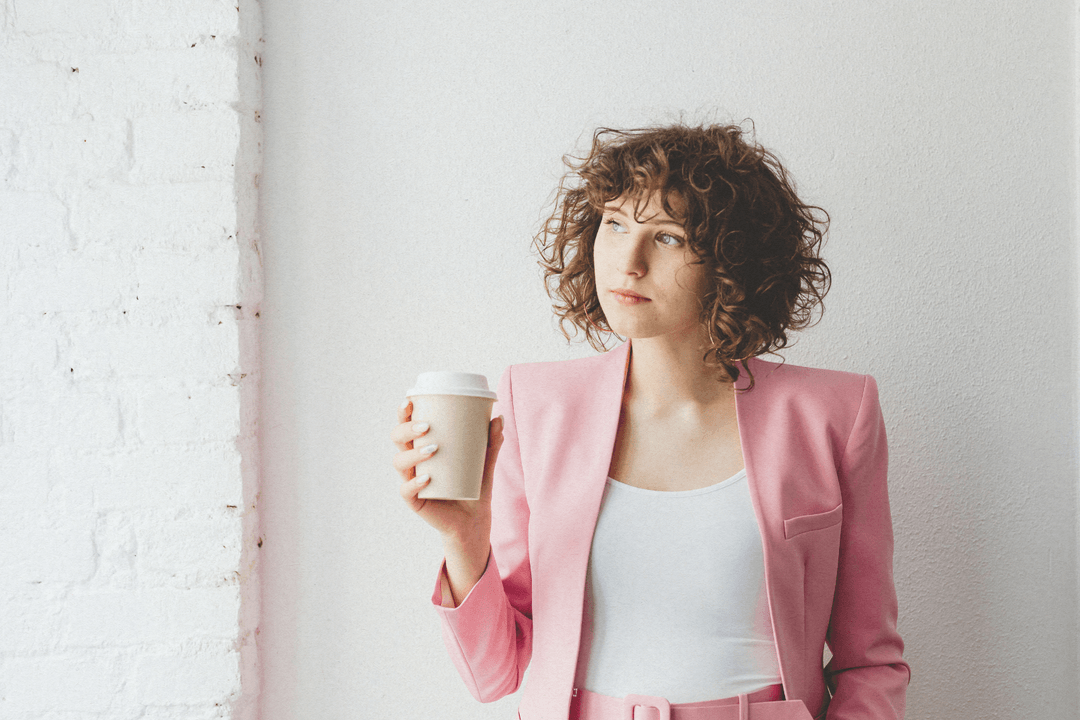 Young woman with curly hair wearing a pink blazer and holding a takeaway coffee cup looking to the side.