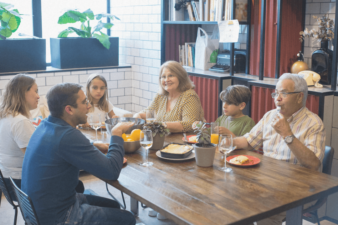 Multi-generational family gathered around a wooden dining table, eating and talking in a bright room with plants and shelves.